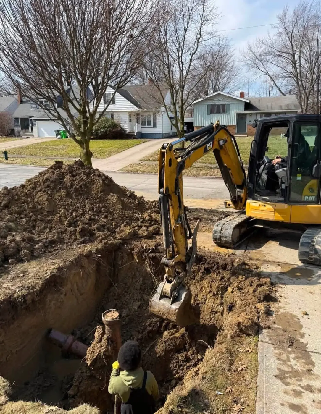 Excavator and worker completing sewer excavation beside a residential street