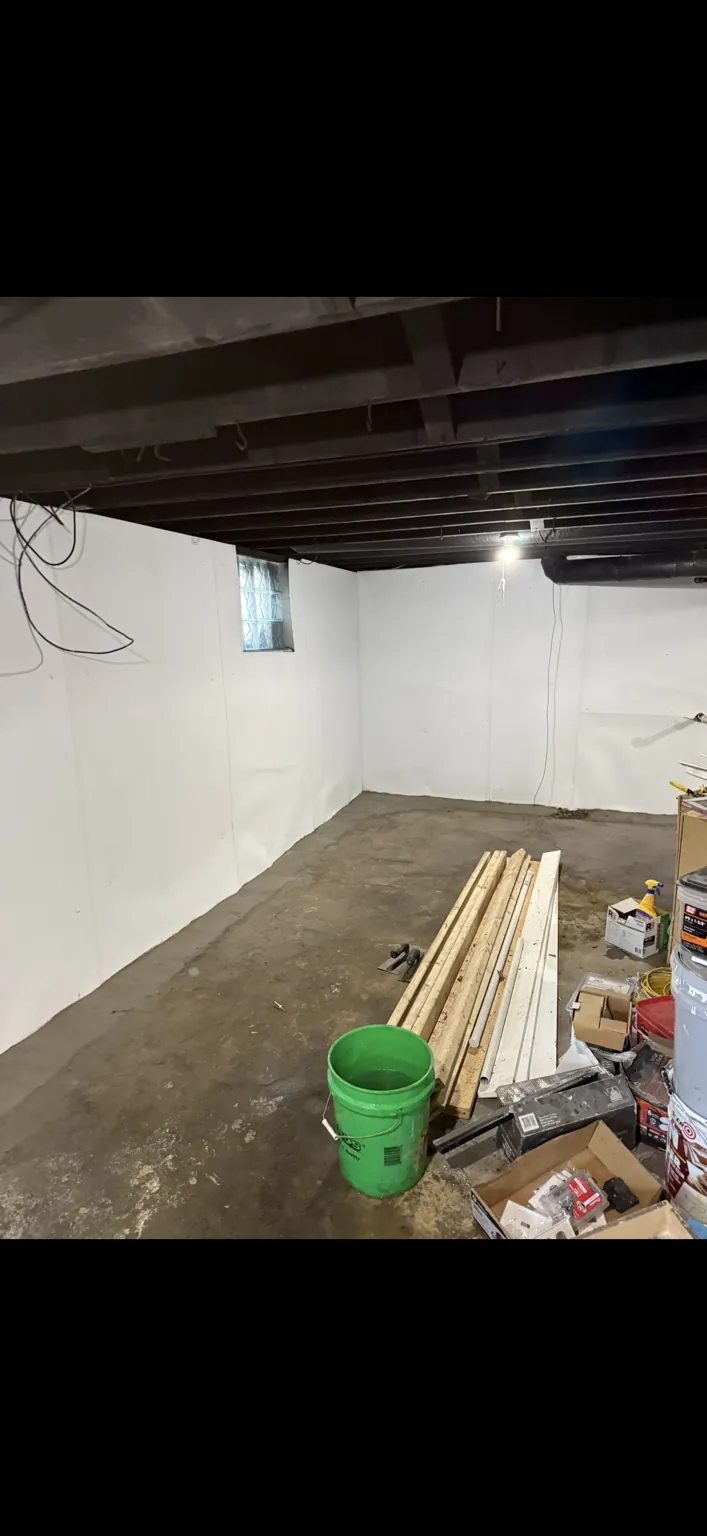 Basement with white wall liner installed along two walls and lumber stacked on the concrete floor