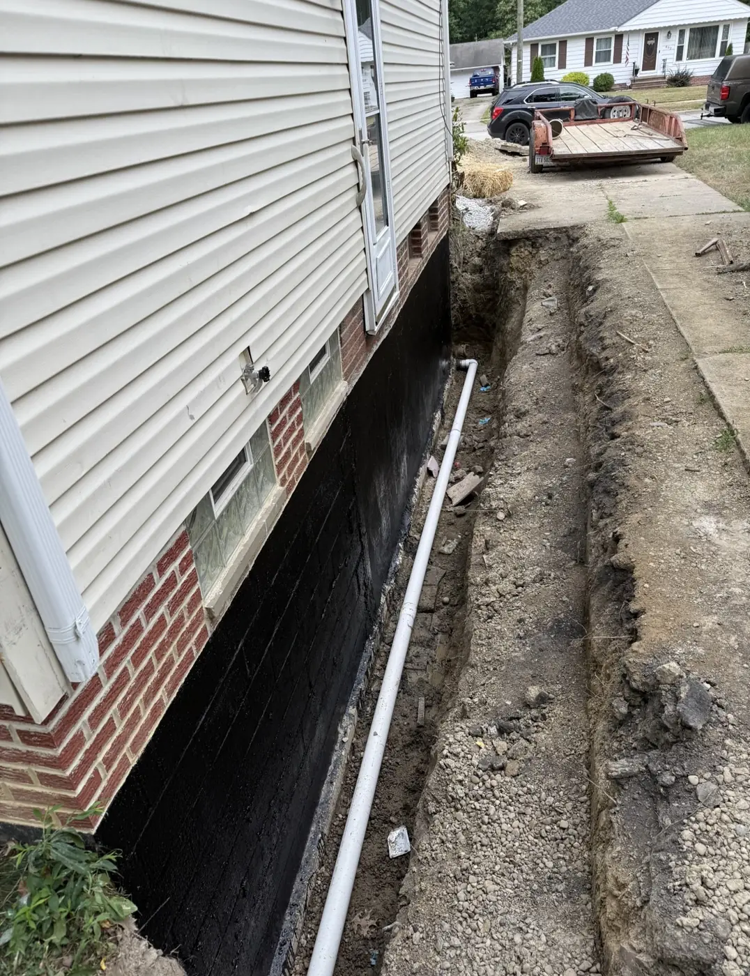 Excavated trench along a house foundation with black waterproof coating applied to the wall and white drain pipe running along the base